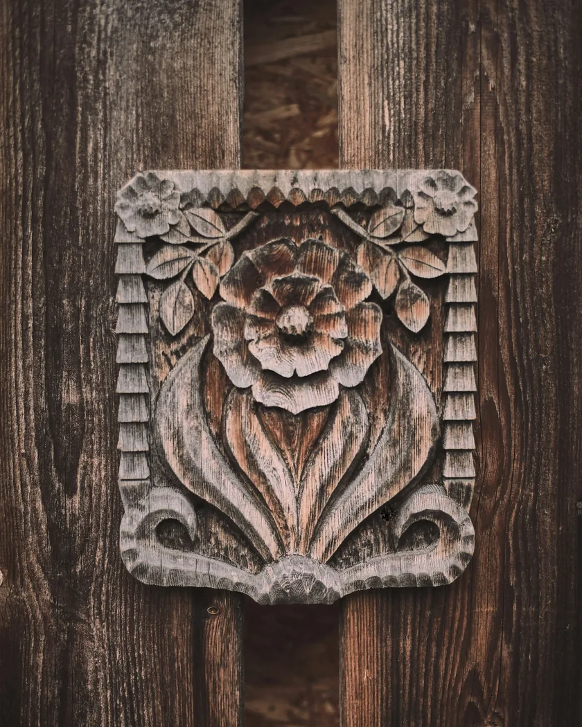 Placa de madera con ornamento floral tallado en relieve, con roseta central, hojas y borde dentado, montada sobre tablas envejecidas