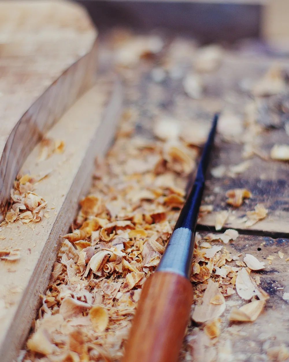 Gubia de mango de madera apoyada sobre un banco de taller cubierto de virutas rizadas junto a una tabla en proceso de talla